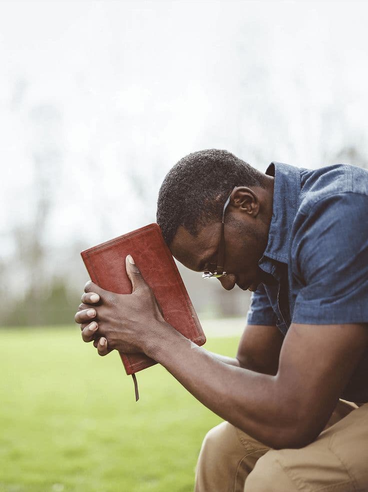 A man kneeling outdoors in prayer, head bowed over his Bible in the morning light